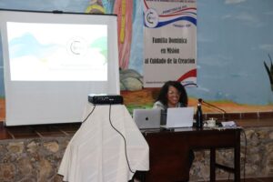 Mujer sentada detrás de un escritorio, junto a una pantalla.